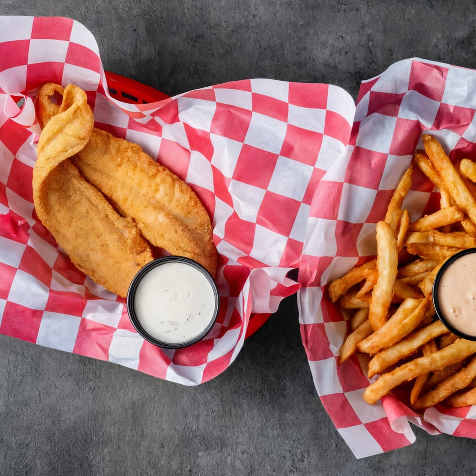 Two baskets of fried fish and french fries with dipping sauces on a dark surface.