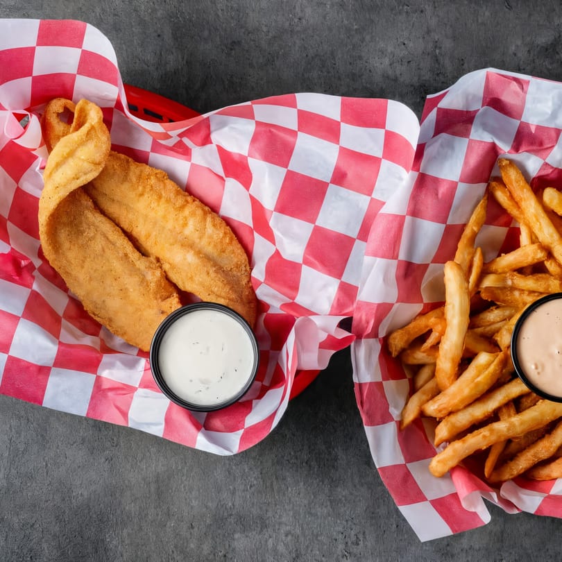 Two baskets of fried fish and french fries with dipping sauces on a dark surface.